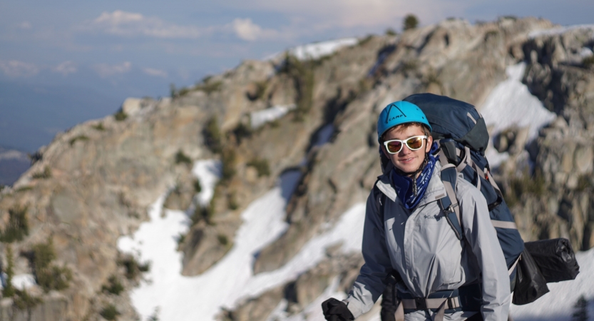 a young person wearing a helmet and a backpacking smiles at the camera. There is a snowy and rocky landscape in the background.